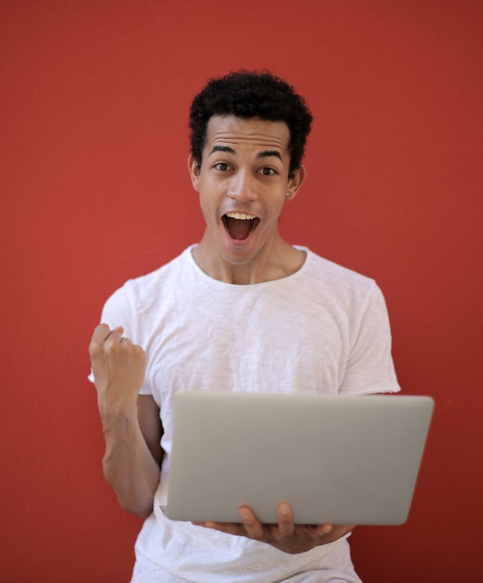 Young man in white shirt with laptop, celebrating success against a vibrant red background.