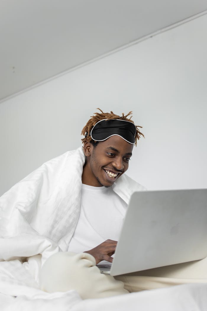 A smiling young man sitting in bed, engaging with a laptop, creating a cozy and relaxed atmosphere.